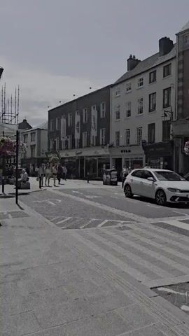 This video captures a sunny day on a bustling street in Kilkenny, Ireland. The scene is a pedestrianized area with shops and buildings lining both sides of the street. In the foreground, a paved walkway is visible, with a trash can on the left. Further down the street, several people are walking, some in small groups, suggesting a leisurely afternoon. The architecture of the buildings appears historic, with some featuring traditional shopfronts. A prominent feature is a clock tower that dominates the skyline, indicating a central town location. Scaffolding is visible around part of the clock tower, suggesting ongoing maintenance or renovation.

The weather appears to be pleasant, with clear skies and bright sunlight casting shadows, implying it is daytime. The mood is calm and ordinary, depicting everyday life in a town center. Some of the shops have visible signage, although the text is mostly unreadable due to the video resolution and distance. One sign, however, appears to read "VILA" on a building to the right. There is a white car driving down the street on the right side of the frame. The overall impression is of a lively and picturesque European town.