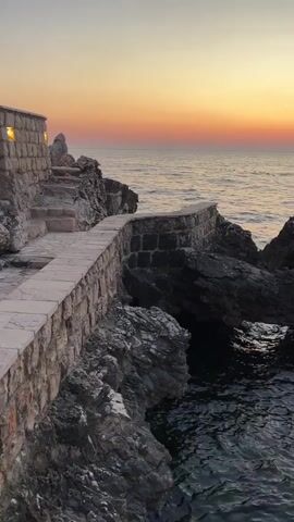 The video shows a view of the sea and rocky coastline at sunset. The sun is setting on the horizon, casting a warm orange and yellow glow across the sky and water. In the foreground, there is a stone pathway and wall leading along the edge of the rocks. The water is dark and reflects the colors of the sky. The rocks are rough and jagged, with some appearing to form natural arches. In the distance, mountains can be seen across the sea. The scene is peaceful and serene, evoking a sense of natural beauty.
