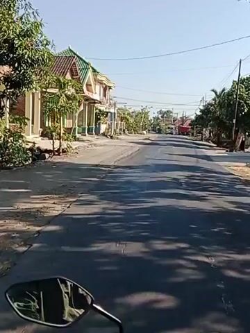 The video captures a street view, likely from the perspective of someone riding a motorcycle, given the presence of a black motorcycle mirror in the foreground. The scene is a rural or semi-rural area with a paved road that appears to be in moderate condition, showing signs of wear and tear. On the left side of the road, there are buildings that resemble houses or small shops, characterized by multiple stories and light-colored facades with green roofs. Vegetation, including trees and bushes, is visible alongside the road and around the buildings. The right side of the road features more trees and what seem to be residential structures in the distance. The sky is clear and blue, indicating a sunny day. In the middle distance, a yellow truck is driving away from the camera. The overall impression is that of a daytime scene on a relatively quiet road with a few signs of life and vehicle traffic.
