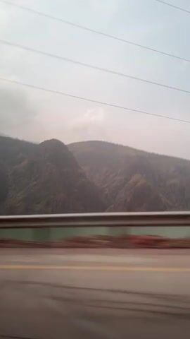The video, taken from the inside of a moving vehicle, shows a landscape featuring mountains and a body of water. The mountains are brownish in color with patches of green vegetation. The water appears greenish. A guardrail is visible in the foreground, partially obscuring the view of the water. The sky is overcast with light grey clouds. The video is shot through a window, as evidenced by a reflection of a wire. The camera remains mostly stable, though the movement of the vehicle causes a blur in the foreground.