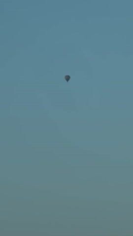The video opens with a close-up of a blurred dark object against a light blue sky, which then transitions to a wider shot of the same sky, revealing a small, colorful hot air balloon ascending. The balloon is the sole object of focus in the sky, appearing in the upper left quadrant of the frame. As the video progresses, the hot air balloon remains visible, gradually moving slightly to the right. Towards the end of the clip, the bottom portion of the frame shows the tops of trees with dark green foliage, indicating an outdoor setting. The lighting suggests it might be early morning or late afternoon. There are no discernible people or interactions within the video.