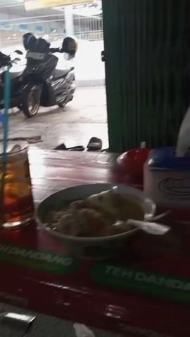 The video opens on a table with a red tablecloth. There are several objects on the table including a bowl of food, a tissue box, a bottle of sauce, and a glass of tea. In the background, a young boy is sitting at the table eating food with a fork and spoon. He is looking at the camera with a serious expression. The scene is dimly lit, and it appears to be an outdoor setting, possibly a street food stall. The camera pans across the table and then focuses on the boy. The video ends with a shot of the table again.
