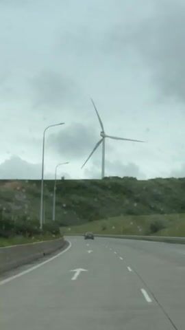 The video depicts a car driving on a highway. Through the windshield, raindrops are visible. On the right side of the road, there is a grassy hill. Atop the hill, a wind turbine stands prominently. In the distance, another car can be seen traveling in the same direction. The sky is overcast with gray clouds. Street lights line the side of the road as well. An arrow marking points left in the driving lane.