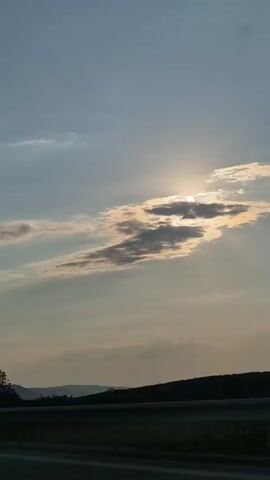 This video captures a serene sky scene with the sun partially obscured by elongated clouds. The sun's rays create a warm, golden glow around the cloud formations, suggesting it is either sunrise or sunset. The sky is a gradient of pale blue and grey, with thinner wisps of clouds scattered across the upper portion of the frame. Below the sky, a dark, silhouetted line of trees marks the horizon, indicating a natural landscape. A dark horizontal line across the bottom of the frame suggests a road or barrier, implying the footage was captured from a moving vehicle or a vantage point at ground level looking across a landscape. The overall mood is peaceful and contemplative, evoking the natural beauty of the sky during twilight. There is no visible text in the video. The context provided places this scene in Brestnitsa, Bulgaria.