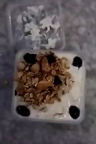 A close-up overhead shot shows a small, square, clear plastic container holding a white, creamy substance, likely yogurt, topped with a pile of granola, scattered raisins, and several cashews.  Around the perimeter of the white yogurt are four evenly spaced dark, round objects that look like blueberries.  To the top left of the main container is a smaller, open clear plastic container holding what looks like small white animal-shaped candies or objects.  The surface beneath the containers appears to be a mottled gray.