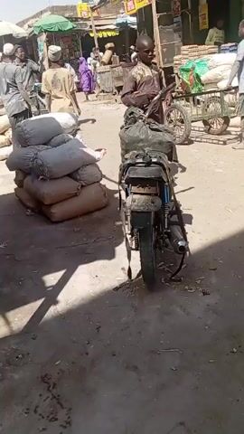 The video shows a busy outdoor market scene, likely in an African country, during the daytime. The ground is dusty and partially covered with debris. Several people are walking around, some carrying items. A motorcycle with bags strapped to the back is parked in the foreground.  To the left are stacks of large white sacks, possibly containing goods for sale or transport. A wooden structure with wire mesh is also visible nearby. In the background, more people can be seen interacting in front of market stalls under awnings. The overall atmosphere is active and commercial.