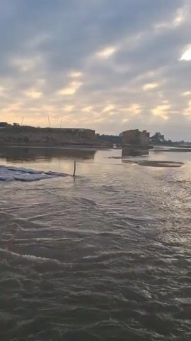 A medium shot captures a river flowing towards the viewer under a cloudy sky with patches of sunlight. The water is slightly choppy. On the left, a man stands on a low, foam-covered structure in the water. A thin stick protrudes from the water near the center of the frame. Across the river, the far bank features a raised area with machinery, possibly a bulldozer, silhouetted against the sky. Debris from a partially destroyed bridge is visible in the river. The overall tone is serene but also shows signs of construction and potential environmental changes.