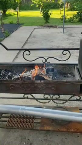 The video shows a close-up view of a metal grill filled with burning firewood. The flames are a vibrant orange and yellow, casting a warm glow on the surrounding charcoal and ash. The grill itself is ornate, with decorative wrought iron scrollwork on its sides.

In the background, a lush green lawn is visible, dotted with trees and some garden lights. The setting appears to be an outdoor space, possibly a backyard or garden in a suburban area. The sunlight suggests it is daytime, and the greenery indicates pleasant weather.

There are no people visible in the frame. The primary focus is the fire in the grill, suggesting an activity like preparing for a barbecue or simply enjoying a fire outdoors. The overall mood is tranquil and evokes a sense of calm and warmth.

No text is visible in the video. The location context provided is Vёшки, Russia.
