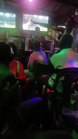 The video shows a group of people in a dimly lit bar, watching sports on two large televisions mounted above the bar. The lighting is a combination of colored lights (red, blue, pink, and green) that create a vibrant but dark atmosphere. Most of the individuals are seated on chairs and stools, facing the televisions. One man, wearing a white tank top and a hat, is standing and moving, possibly dancing or reacting to the game. Other people are visible only from the back and sides. The environment appears lively, with sounds of music or commentary mixing in the background. The overall setting suggests a social gathering to watch a sporting event in a casual bar.