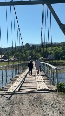 Two individuals walk across a suspension bridge made of wood. The bridge is supported by cables and has metal railings. A river flows beneath the bridge. In the background, there are houses and trees on a hill under a clear blue sky. The bridge appears weathered, with some wooden planks out of place. One person is wearing a dark jacket and pants, while the other wears a striped shirt and shorts and carries a bag.