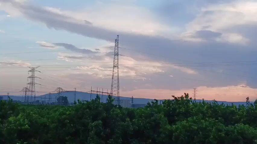 The video pans across a rural landscape at dusk. In the foreground, a dense grove of green trees, likely citrus, stretches across the frame. Beyond the trees, a series of tall, lattice-like electricity pylons stand silhouetted against a cloudy sky painted with soft hues of pink and blue. Further in the distance, rolling hills dotted with scattered houses and the faint outlines of more power lines create a sense of settlement within the natural environment. The overall atmosphere is tranquil and serene, capturing the transition from day to night in a power-generating region.