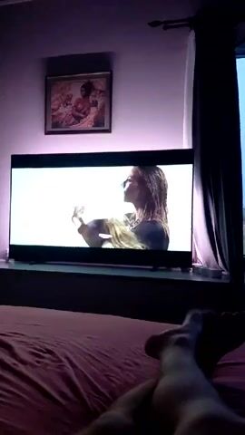 A first-person point of view shot from a bed shows a large television on a stand against a wall displaying various clips from what appears to be a music video. The television is illuminated with backlighting that changes colors to match the video's content. A framed picture hangs above the television. A window with a curtain on its right is visible. A person's bare feet are visible in the foreground.