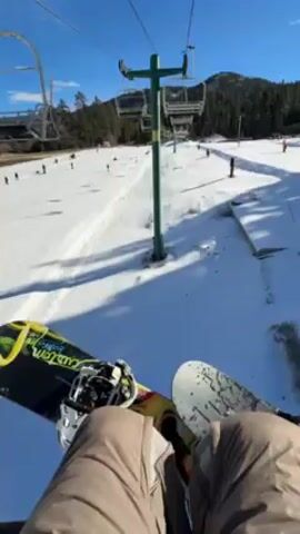 The video shows a first-person point of view of someone on a ski lift, holding a snowboard between their legs. The lift passes over a snowy hill with scattered pine trees and a few people skiing and snowboarding. The camera then pans up to show the person riding the lift, wearing a flannel shirt, and another person next to them wearing a helmet and goggles. Both people give a peace sign to the camera. The sky is bright blue and clear.