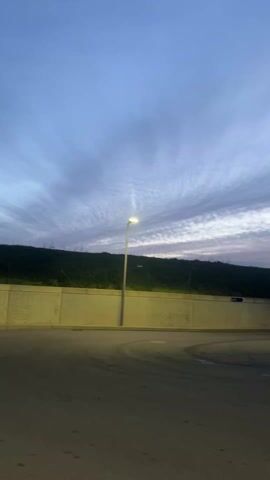 The video depicts a curved section of asphalt road under an early morning or late evening sky with cirrus clouds streaked across the horizon. A tall concrete wall stands adjacent to the road, topped with a green hillside and a wire fence. A streetlamp casts a warm glow on the road and the wall. A green truck slowly drives into the frame from the right.