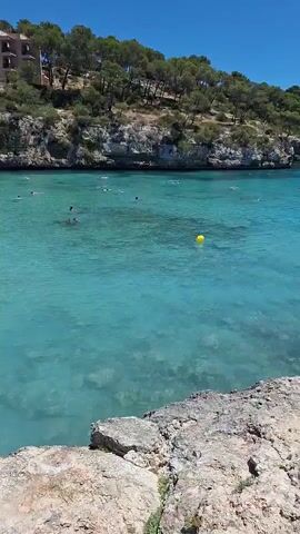 The video captures a scenic view of a bay with clear turquoise water. The water is dotted with yellow buoys. Several people are visible swimming and enjoying the water. In the distance, a few boats are anchored. The bay is surrounded by a lush, green, tree-covered hillside on one side and rocky terrain on the other, with a pathway visible on the right. The sky is clear and blue. The camera pans slightly to the right, revealing more of the bay and the surrounding landscape.