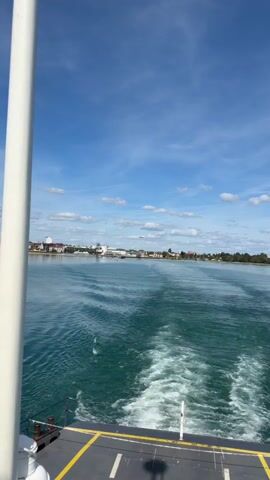 The video shows a view from a moving boat on a body of water. The boat is leaving a wake behind it. In the distance, there is a shoreline with buildings and trees. Above the shoreline, a blimp is visible in the sky. The sky is blue with some white clouds.