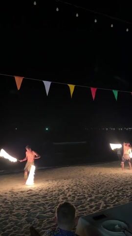 The scene depicts two men performing a fire dance on a sandy beach at night. Both men are shirtless and wearing some sort of skirt or wrap around their waist. They each hold flaming batons and twirl them in a coordinated manner. The beach is illuminated by the firelight and overhead string lights. In the foreground, a person is sitting at a table, partially obscured.  The background shows the dark expanse of the ocean and a distant, faint city skyline. Colorful triangular flags are strung overhead.