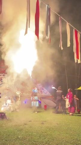 A performer dressed in a blue and red costume with a feathered headdress stands in front of a large tent decorated with red and white fabric strips hanging from above. The performer is holding a device that is expelling a large jet of flame and smoke upwards. To the left of the performer, a small pile of burning material is visible on the grass. Several people are standing to the right of the performer, observing the display. The scene appears to be taking place outdoors at night, with trees visible in the background.