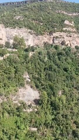 The video pans across a mountainous landscape covered in lush green trees. Sheer rock cliffs rise from the dense foliage, with layers of stratified rock visible. A narrow path or road winds along the cliff face in the upper portion of the frame. In the lower right quadrant, a railing of dark metal bars with wooden accents is visible, suggesting a viewpoint or walkway. A multi-story building with a light-colored facade and windows is nestled into the cliffside, with trees growing around it. The sky above is a clear, bright blue.
