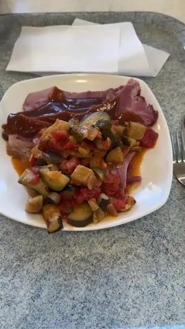 A plate of food sits on a speckled gray countertop. The plate is white and contains several slices of ham covered in a dark brown sauce, topped with a colorful mixture of chopped vegetables including zucchini, tomatoes, and eggplant. A fork and knife are visible on the right side of the plate. A white napkin is in the background. A person's hands are cutting into the ham with a knife and fork. The scene is lit from above.