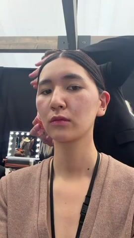 A young woman with dark hair pulled back and no makeup is being prepped for a fashion show. She wears a heather tan cardigan and has a black lanyard around her neck. A woman with long blonde hair and a black jacket is smoothing the first woman's hair. There are other models and stylists in the background. The lighting is bright and the setting appears to be backstage at a fashion event.