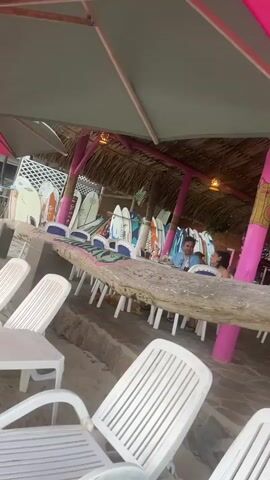 The video shows the interior of a beachside structure with a thatched roof and pink support beams. Below the roof are decorative lights and hanging ornaments. Tables and chairs are arranged throughout the space. There are people sitting at one of the tables. White plastic chairs are visible in the foreground. The camera angle tilts upwards, focusing on the roof and decorations.