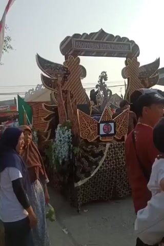 The video shows a parade float decorated with intricate batik patterns. The float is adorned with traditional Indonesian motifs, including what appear to be wayang kulit-inspired cutouts and a sign that reads "PAGUYUBAN BATIK PUNGGARI". A large number "51" is prominently displayed. Several people are visible on and around the float, some seated and others standing. The float is moving along a street, suggesting a parade or a festive procession. The overall atmosphere appears to be bright and outdoors, with a clear sky.