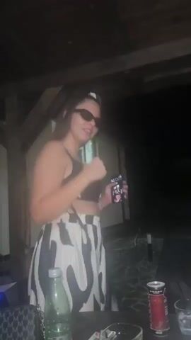 A young woman with long dark hair, wearing sunglasses, a black crop top, and black and white patterned wide-leg pants, stands under a wooden structure at night. She is dancing, smiling, adjusting her sunglasses, and touching her top. On a table in the foreground, there is a green glass bottle, a red can, a black bag, and two glasses. The background is dark with glimpses of a building.