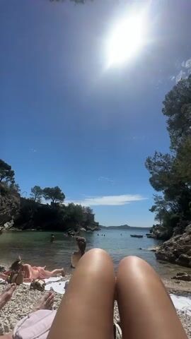 The video shows a first-person view of someone lying on a beach. The person is wearing light-colored clothing, and their legs are visible in the foreground. In the background, there's a small beach cove with clear blue water and a bright sun shining overhead. There are other people on the beach, some sunbathing on towels and others swimming in the water. The beach is surrounded by rocky cliffs and trees on either side, creating a secluded and picturesque scene. A small boat is anchored in the distance. The sky is clear and blue with a few clouds visible near the horizon.