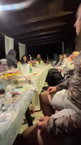 A group of people are gathered around a long table at night. The table is covered with a white tablecloth and laden with food and drinks, including bottles of alcohol, glasses, and plates of food. There is an ashtray on the table. The people appear to be socializing and enjoying a meal together. The scene is lit by overhead lighting, casting a warm glow. The background suggests an outdoor setting, possibly a patio or porch, with a wooden ceiling and white curtains. Some individuals are looking at the camera while others are engaged in conversation or eating. The overall atmosphere seems relaxed and convivial.