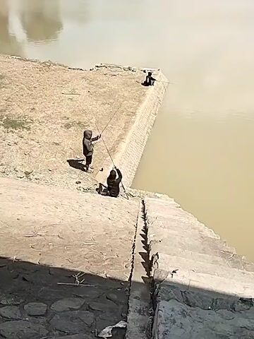 The video shows three people fishing by a body of water. Two people are closer to the camera, one standing and one sitting, both holding fishing rods. A third person is further away, also fishing. The area around the water appears to be dry and dusty, with some concrete structures along the edge of the water. The water itself is a murky brown color. The sky is clear and the lighting suggests it is daytime.