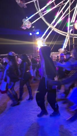 The video shows a nighttime scene at what appears to be a festival or carnival. A large Ferris wheel illuminated with various colors of light dominates the background.  People are walking through a sandy or dirt-covered area.  Several individuals are clearly visible, including one wearing a black hoodie with the hood up and black pants, and another individual in a dark blue t-shirt.  The lighting is predominantly blue and purple, which makes it difficult to discern specific details of the surrounding environment, but there appear to be food stands or other vendor structures in the background.  The camera appears to be handheld and moves, capturing the motion of people walking by.