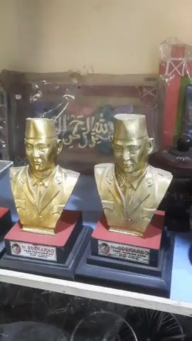 The video pans across a shelf displaying several gold-colored busts and full-body statues of a person, likely a historical figure given the name "SOEKARNO" visible on some of the bases. Two busts are prominently featured in the foreground, followed by more busts and statues encased in glass display boxes. In the background, there are various items, including what appears to be framed artwork with Arabic calligraphy and other objects covered in plastic wrap. The lighting suggests an indoor setting, possibly a shop or display area. The video is shaky at times.
