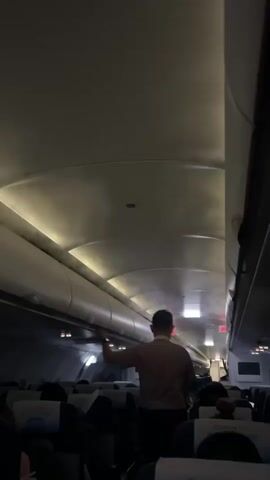 The video shows the interior of an airplane cabin from a low angle, looking towards the front. The lighting is dim, with overhead lights illuminating the curved ceiling and air vents. A flight attendant, wearing a light-colored shirt and dark pants with a cap, is standing in the aisle and spraying a fine mist from a handheld device towards the overhead compartments. Passengers are seated in dark-colored seats with blue headrests visible in the foreground and background. The aisle appears to be empty except for the flight attendant. The video is short and appears to capture a moment during a safety or comfort procedure.