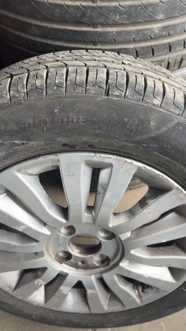 The video shows a close-up of a tire with a screw embedded in its tread. A clear liquid, likely soapy water, is sprayed onto the tire around the screw. Air bubbles are visible, indicating that air is leaking from the tire around the screw. The tire is mounted on a rim, and other tires are visible in the background, stacked on top. The lighting is somewhat dim, and the tires appear used and slightly dirty.
