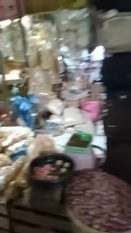 The video shows a busy outdoor market scene. There are numerous stalls selling various goods, including clothing, food items, and possibly crafts. People are seen walking through the market, browsing the stalls, and interacting with vendors. The environment appears to be a covered walkway or a somewhat enclosed market with wooden structures and a dirt or concrete floor. The lighting is dim, suggesting it might be early morning or late afternoon. Some stalls have goods displayed on wooden pallets or tables. The overall atmosphere is one of bustling commerce and everyday life in a market setting.