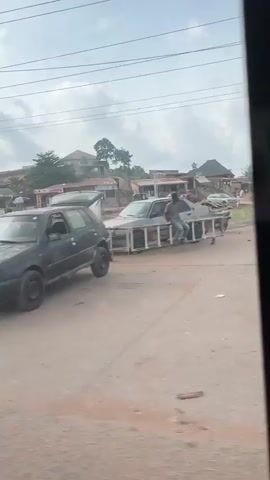 The video shows a street scene in a developing area, likely in Africa. The road is dirt, with several parked cars including a silver sedan, a pink sedan, a white van, and a blue SUV. Pedestrians are walking along the street, some dressed in traditional patterned clothing. There are several small shops or stalls along the side of the road, with corrugated metal roofs. The sky is cloudy, and power lines run along the top of the frame. In the distance, a hill or elevated area can be seen, with buildings on top. A large truck is parked further down the road.