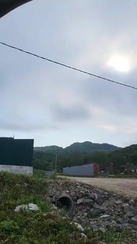 A video shows a cloudy sky with the sun visible through the clouds. There are hills covered in trees in the background. In the foreground, there is a large green building on the left, a dirt road, and a concrete pipe with rocks around it. A power pole with wires is visible in the middle right of the frame.