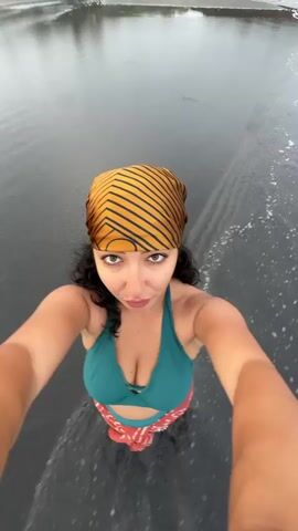 A woman stands in shallow water on a dark-sand beach, recording a selfie video. She has dark curly hair and wears a patterned scarf on her head, a green bikini top, and a patterned sarong around her waist. The water is dark and reflective, with small waves lapping around her feet. The beach is visible in the background, with some vegetation and rock formations. The sky is overcast, creating a slightly diffused light. She looks directly at the camera with a neutral expression.