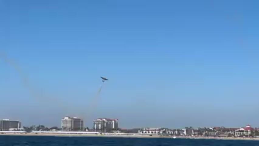 A small airplane ascends, leaving a white smoke trail in a clear blue sky above a coastal city skyline.