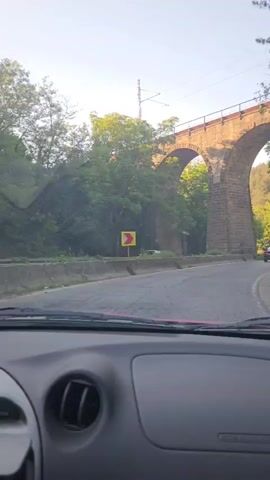 The video shows a view from inside a car driving on a road. A large stone viaduct with arched openings is visible on the left side of the road. Trees and a wooded hillside are on the right side. The sun is shining, casting shadows. Another car is visible ahead on the road.