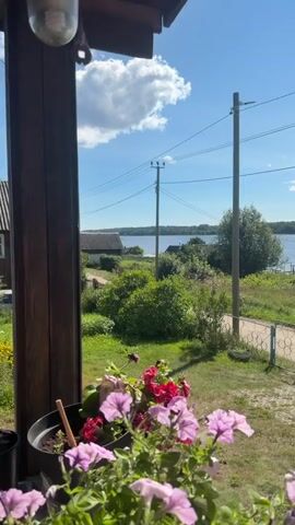 The video shows a view from a porch, looking out at a rural landscape on a sunny day. In the foreground, there are purple and red flowers in a pot. Beyond the porch, there is a grassy yard with a decorative fence and a path. In the middle ground, there are trees and bushes, and a body of water, likely a lake or river, is visible. Several utility poles with wires stretch across the scene. In the background, there are some houses and a treeline on the far side of the water. The sky is blue with some fluffy white clouds. The video is short and appears to be a static shot with no significant movement or action.