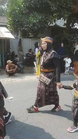 The video shows a procession of people walking down a street. In the foreground, a woman in traditional clothing, including a headscarf and patterned skirt, holds the hand of a child also in traditional attire. The child wears a matching headpiece and patterned outfit. Behind them, other adults and children in similar traditional clothing are visible. Some adults are wearing masks. In the background, there are houses, trees, and parked motorcycles. People are standing on the side of the street, observing the procession. The weather appears to be sunny.