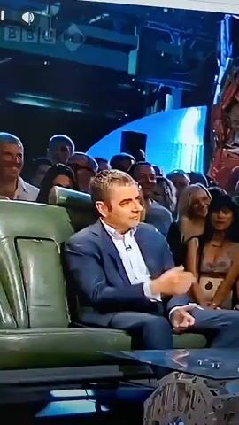 A man in a blue suit and checkered shirt is sitting in a car seat, talking to a crowd. He is gesturing with his hands. Another man in a tan jacket is visible to the right, looking down. The background is blurry, but appears to be a stage with blue lighting and people in the audience. Text overlays appear on the screen: "IT JUST-", "LIKE IT", "OF SAYING IT", "BOB", and "THAT BOB". The video is from BBC.