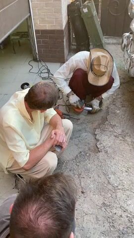 In a courtyard with a concrete floor, three men are gathered around a grinding tool. One man, wearing a light-colored shirt and tan pants, kneels with his hands clasped, observing. Another man, dressed in a white shirt, dark pants, and a straw hat, crouches and operates the grinding tool. A third man, whose back is to the camera, is partially visible in the foreground. The environment appears to be outdoors, with a brick wall in the background and a partially open garage door to the left. The scene is illuminated by natural light.
