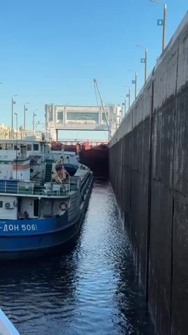 Two boats are moving through a lock. The boat on the left is a smaller blue and white vessel with the number 5061 on its side. The boat on the right is a larger, dark red cargo ship, only its bow is visible. The boats are moving towards a large concrete structure that appears to be a lock gate. The sky is clear and blue, and the water is calm. The scene appears to be in daylight.