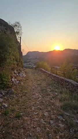 The video begins with a view of a dirt path winding through a natural landscape at sunset. The sun is visible on the horizon, casting a warm glow over rolling hills and mountains in the distance. A dry stone wall with a metal railing runs along the right side of the path, separating it from a grassy area. Lush green vegetation grows on the left side of the path, bordering a rocky cliff face. A single utility pole with wires is also present on the left. The camera pans slightly to the right, offering a wider view of the valley and the setting sun. The path appears to lead into the distance, towards the sun-drenched landscape. The overall scene evokes a sense of tranquility and natural beauty.