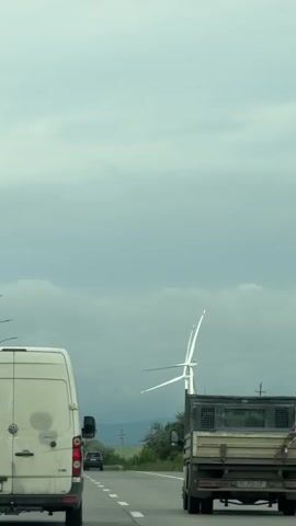 The video depicts a highway scene with a white van as the primary subject.  The van is driving on a multi-lane road, and in the background, a wind turbine is visible. Streetlights line the highway. The sky is overcast.  A truck is driving in front of the van. The sound of music is audible.