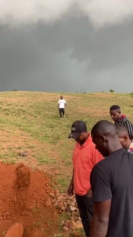 The media file depicts a somber outdoor scene with several individuals gathered on a grassy, reddish-brown hillside. The sky is heavily overcast with dark, dramatic storm clouds, suggesting impending rain or a recent downpour.

In the foreground, three men are visible. Two are facing away from the camera, with one wearing a black t-shirt and the other a black baseball cap and an orange polo shirt. The third man, slightly to the left and further back, is wearing a black t-shirt. In the middle ground, a man in a white t-shirt and dark pants is walking away from the camera up the hill. The ground in the immediate foreground is disturbed, with a pile of reddish-brown soil, possibly indicating a recently dug grave.

The overall mood of the scene is solemn and perhaps a bit unsettling due to the dark weather and the context of the disturbed earth. The location appears to be rural or semi-rural, with open space and a natural landscape. There is no visible text in the media file. The activity taking place is unclear but appears to be related to a burial or memorial service, given the presence of the soil pile and the gathered individuals. The time of day is difficult to ascertain precisely due to the cloud cover, but the overcast conditions suggest it could be daytime, possibly late afternoon.