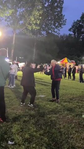 Two men dance on a lawn at dusk. The man on the left wears a dark jacket, dark pants, and black-and-white athletic shoes. The man on the right wears a red shirt under a navy jacket, dark pants, and dark slip-on shoes. They face each other and move their arms and legs to music playing in the background. Behind them, a crowd of people stands or walks by. In the background, there are trees, lights, and a brightly colored carousel. The camera briefly pans left as people walk in front of it.